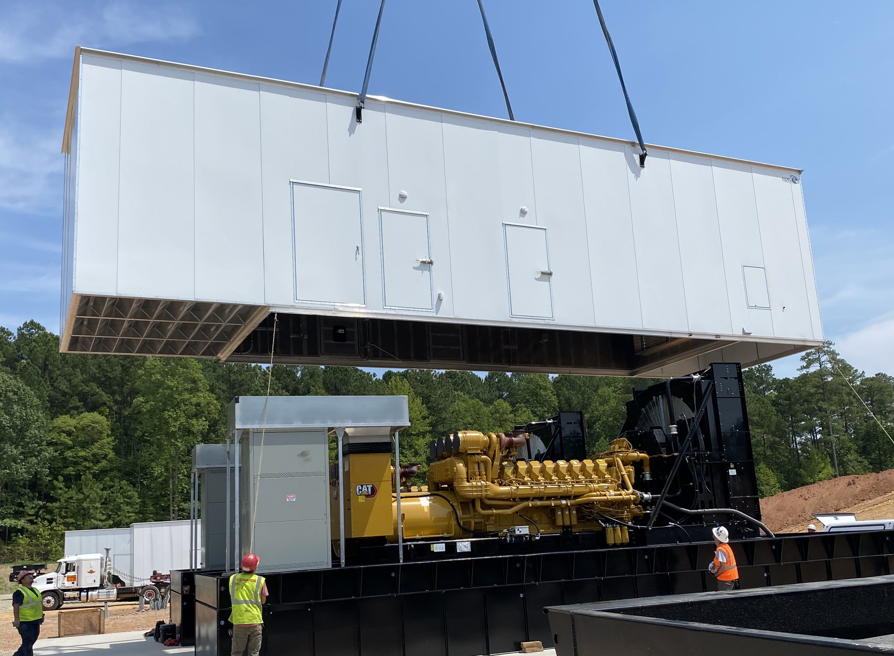 Crane placing white box enclosure over a large, yellow backup generator