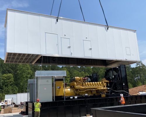 Crane placing white box enclosure over a large, yellow backup generator